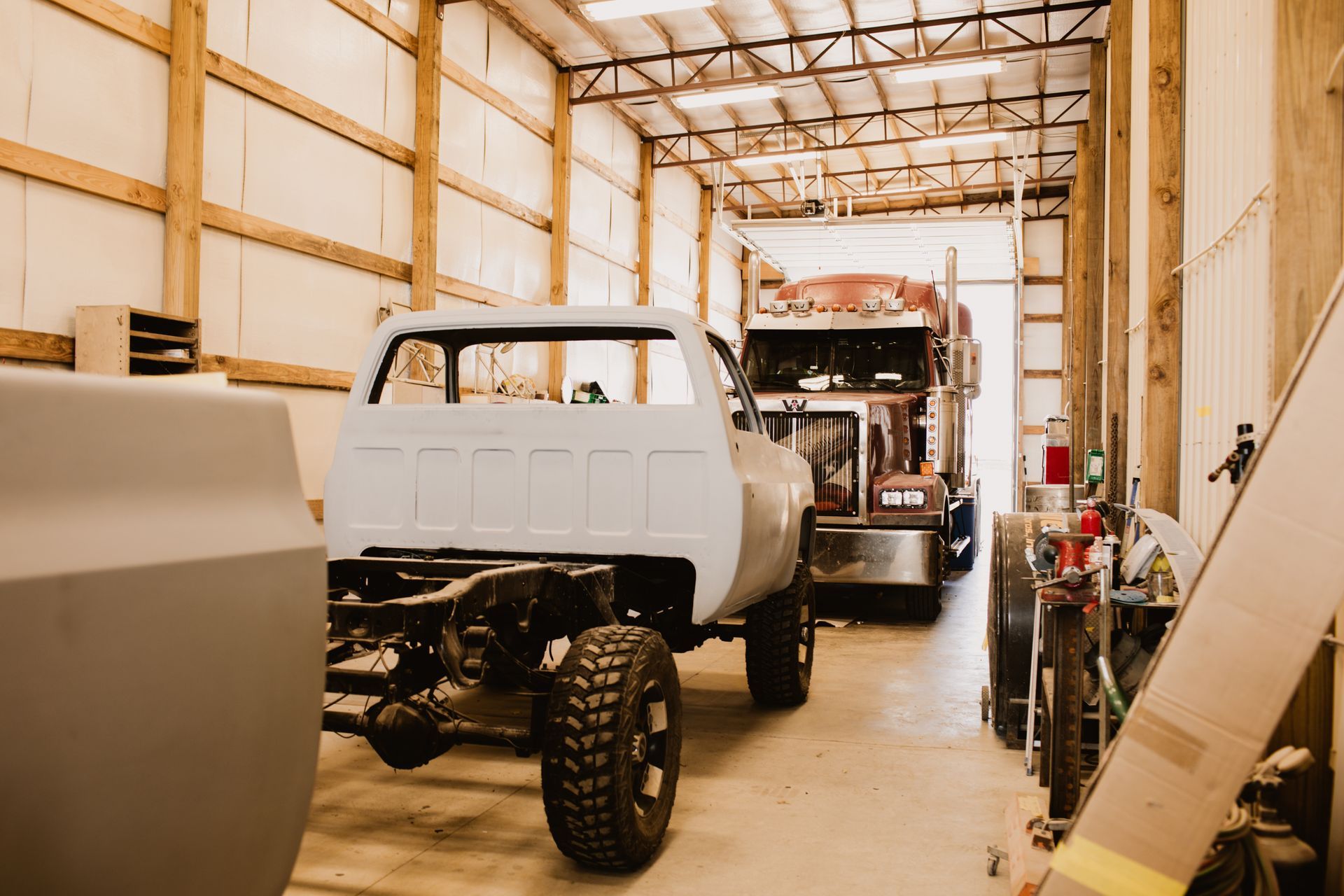 A light-colored pickup truck frame under repair in a workshop, with a large semi-truck parked behind it.