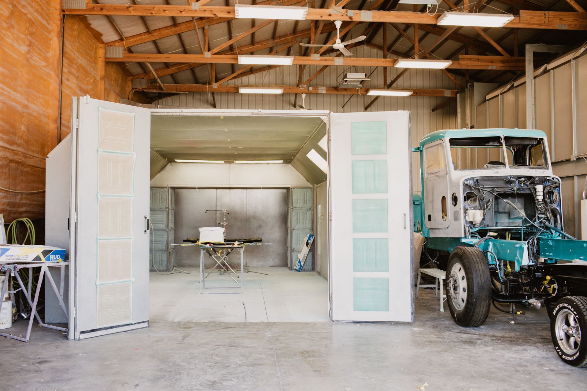 An industrial spray paint booth with open doors stands in a garage next to the chassis and cabin of a disassembled truck.