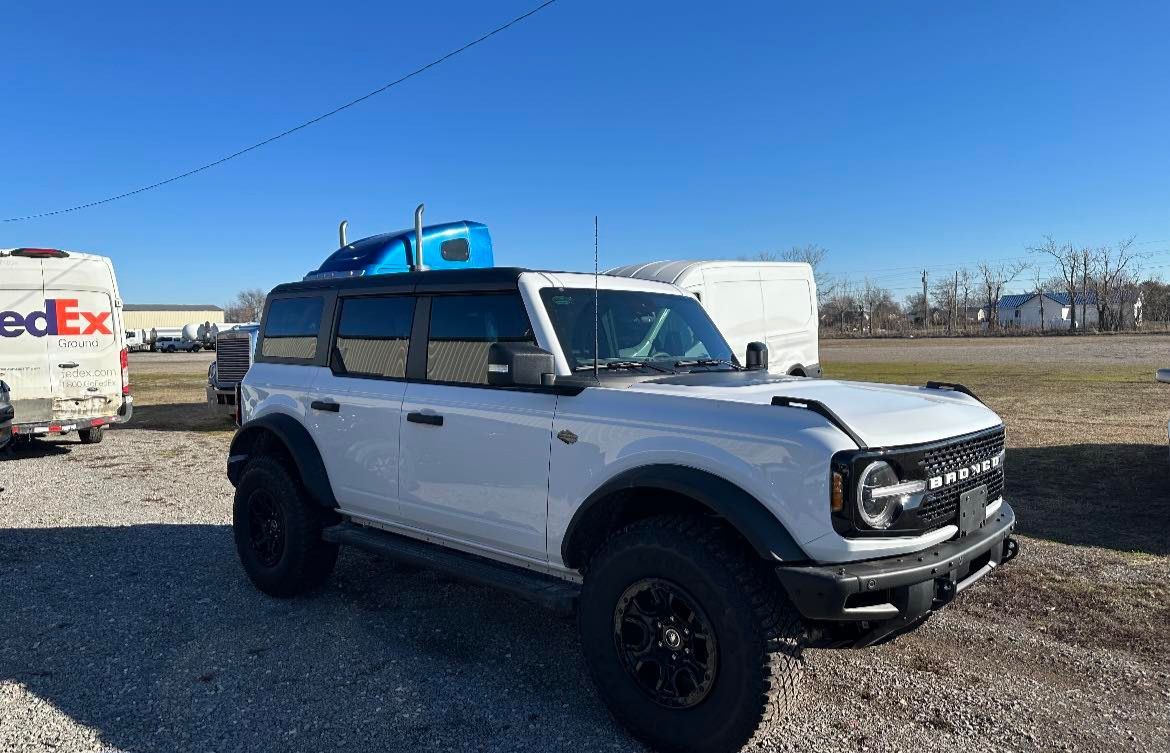 A white Ford Bronco parked on a gravel lot next to a FedEx truck under a clear blue sky.