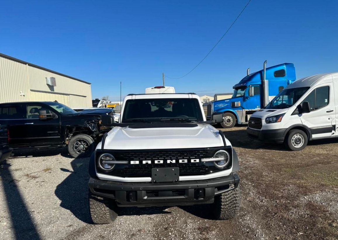 A white Ford Bronco parked outdoors in a gravel lot with a blue semi-truck and white van in the background.
