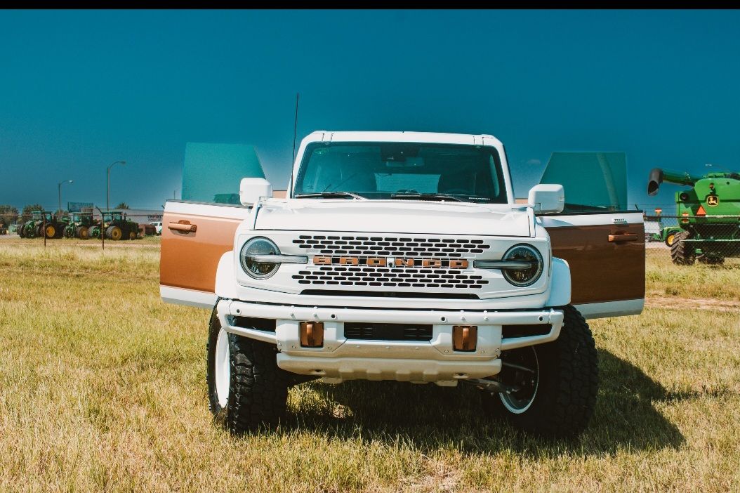 A white Ford Bronco with open doors parked in a grassy field, with farm machinery visible in the background.