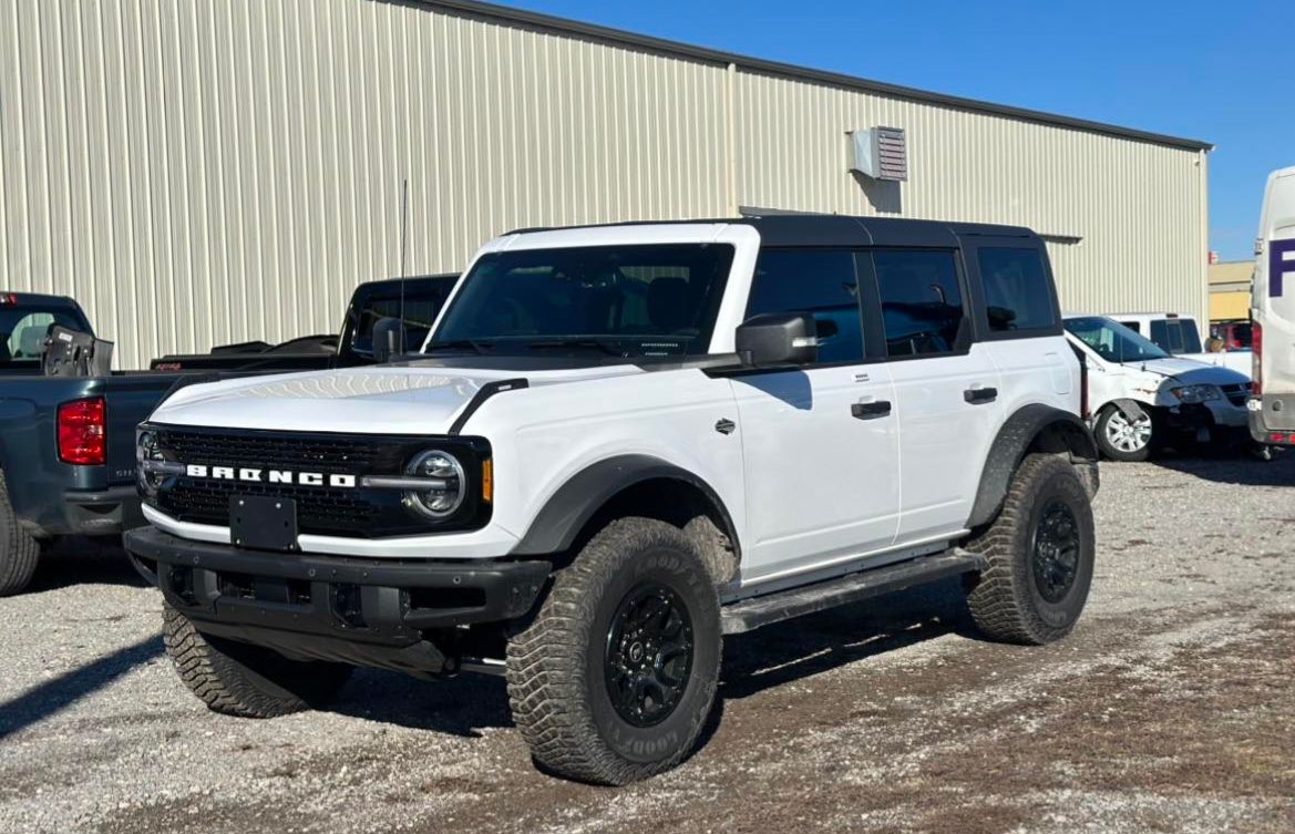 White four-door Ford Bronco parked on a gravel lot in front of a metal warehouse building.