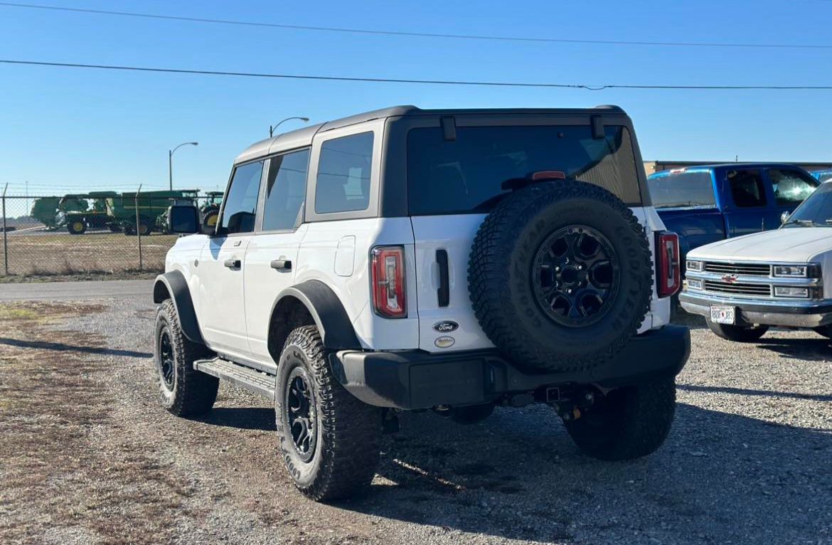 A white Ford Bronco SUV parked on a gravel lot under a clear blue sky, viewed from the rear three-quarter angle.
