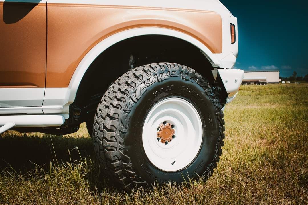 A close-up of a white and copper-colored truck's front wheel, featuring a large off-road tire parked in a grassy field.
