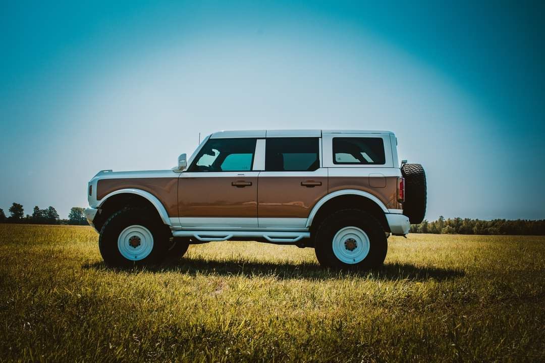 A two-toned tan and white Ford Bronco parked in a grassy field under a bright blue sky.