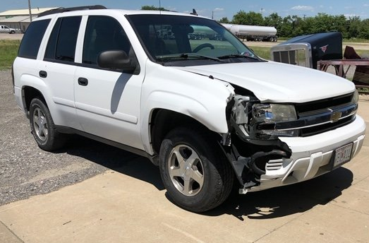 White SUV parked on a paved lot with significant front-end damage to the driver-side bumper and headlight assembly.