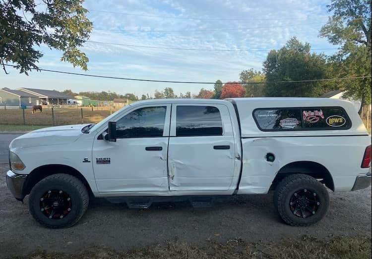 White pickup truck with damage to the side doors and rear quarter panel, parked on a gravel drive near a farm.