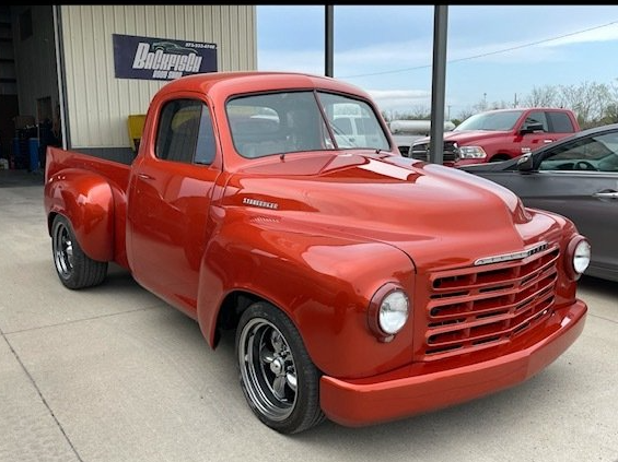 A glossy, metallic-orange vintage Studebaker pickup truck parked on concrete outside an auto shop.
