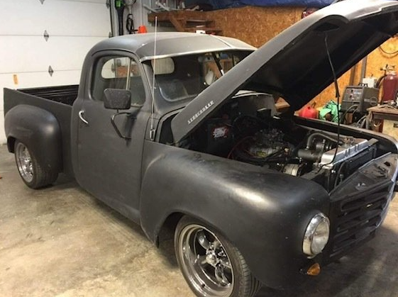 A matte black vintage pickup truck with its hood open, parked inside a garage.