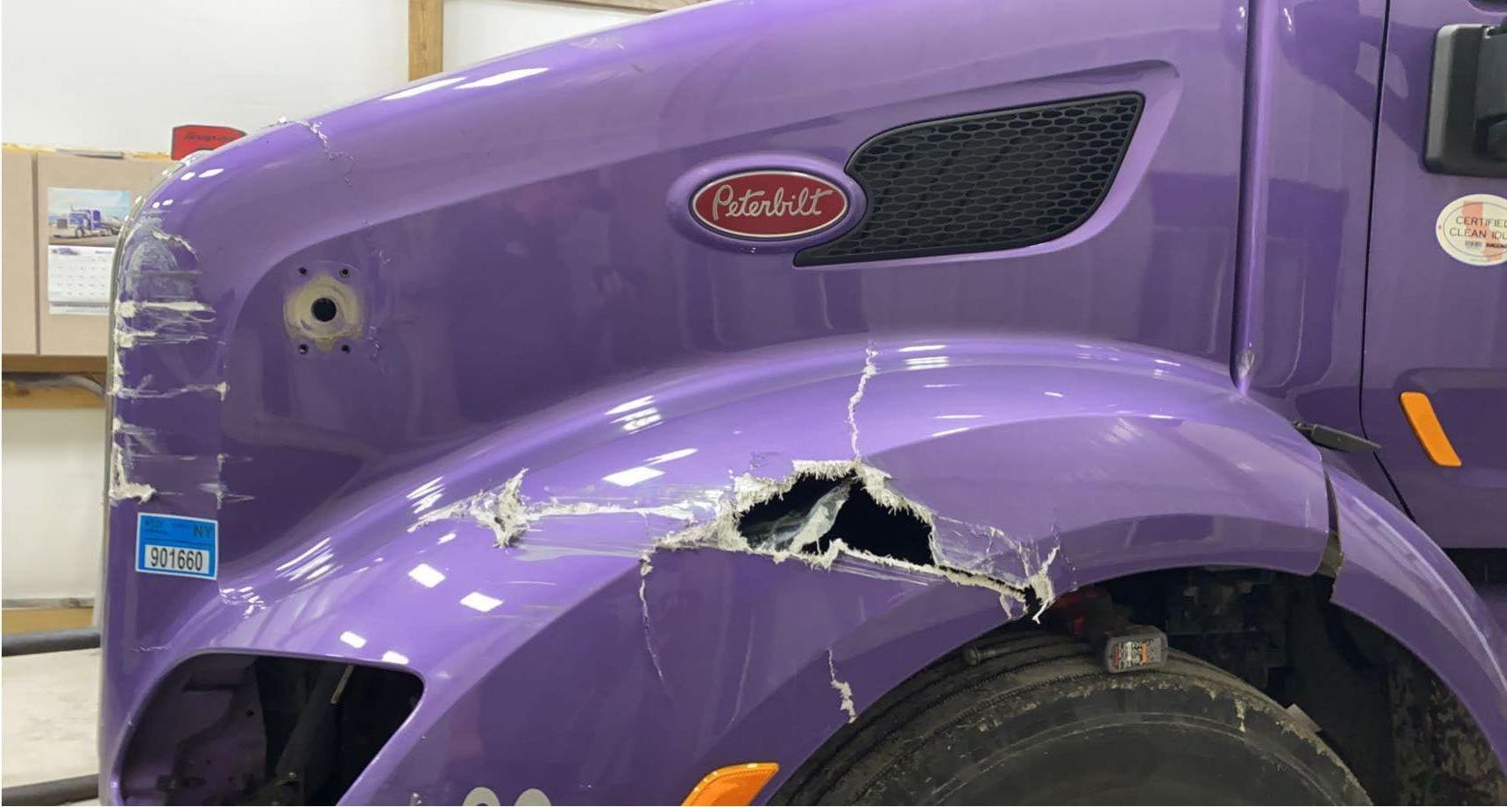 Close-up of a damaged purple Peterbilt truck hood with a large puncture and scratches above the front wheel.