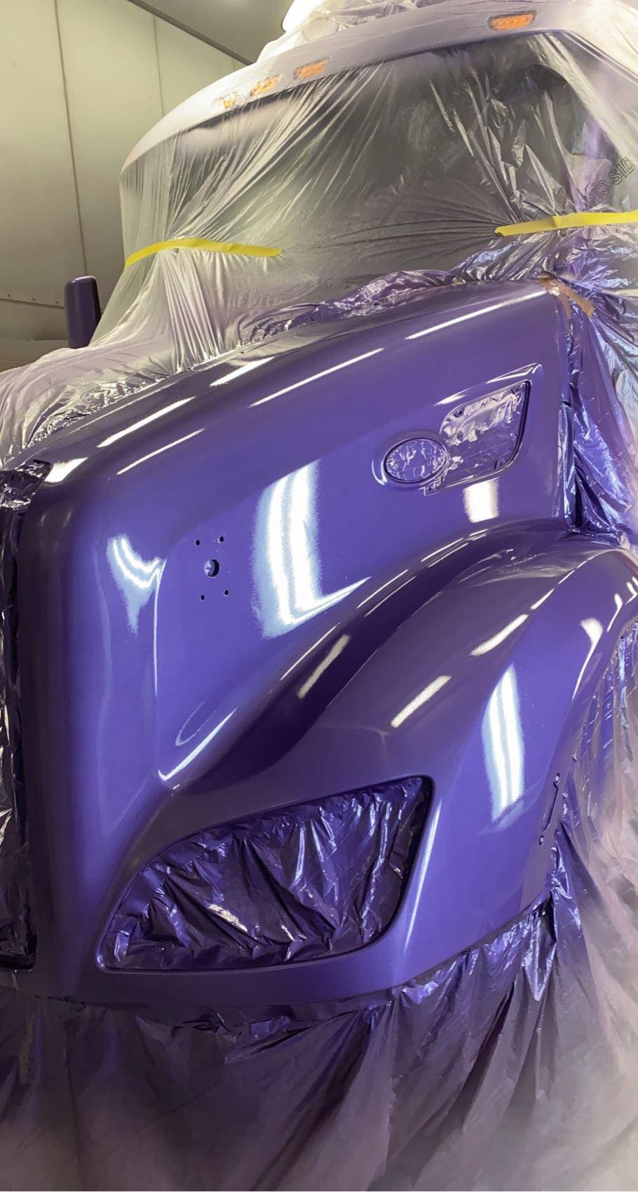A glossy, freshly painted purple semi-truck hood in a spray booth, partially covered in protective plastic sheeting.