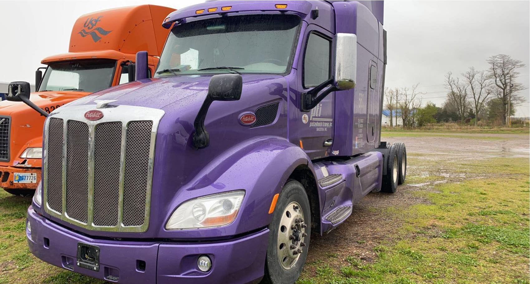 A purple Peterbilt semi-truck parked on a grassy lot next to an orange truck.