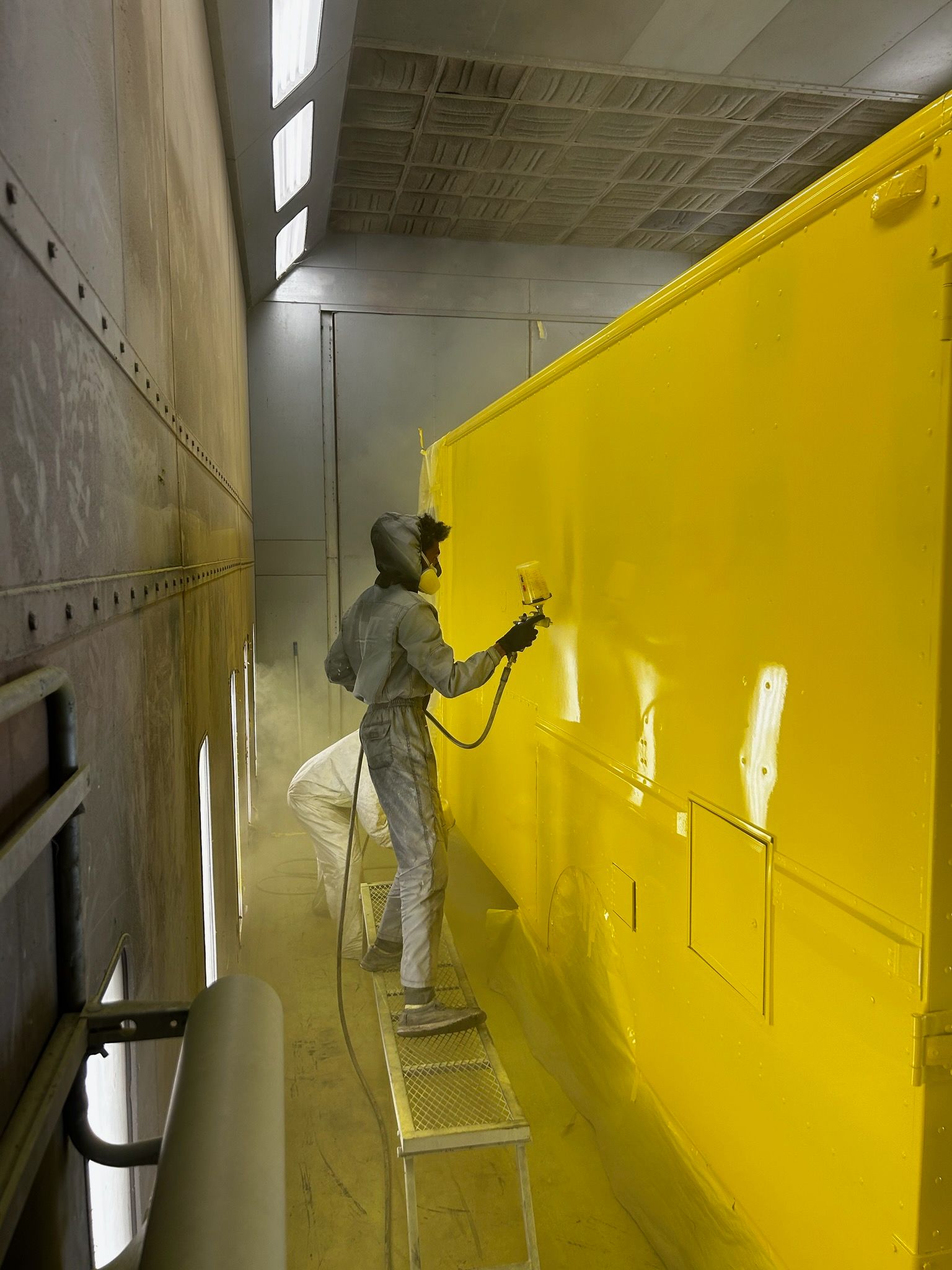 A worker in protective gear uses a spray gun to paint a large metal structure bright yellow inside an industrial booth.