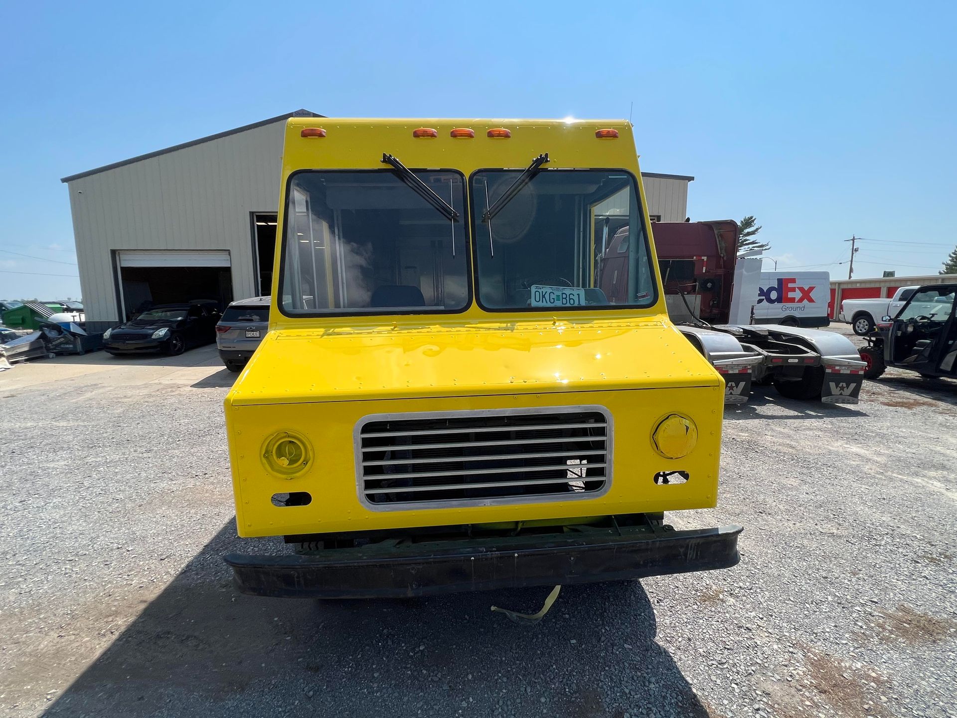 A yellow step van parked on a gravel lot in front of a warehouse on a sunny day.