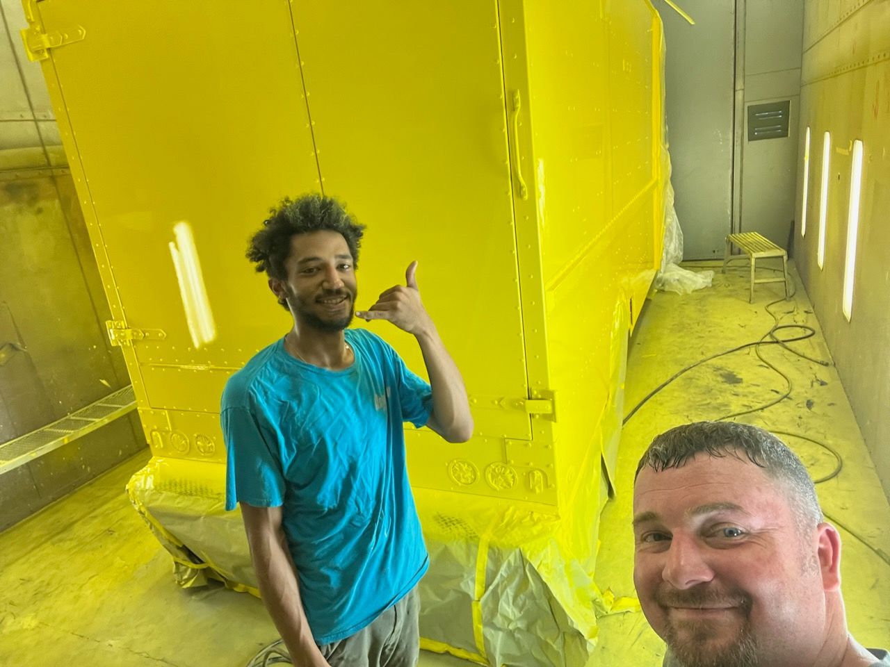 Two people smile inside a workshop, one making a hand gesture, in front of a large, freshly painted bright yellow object.