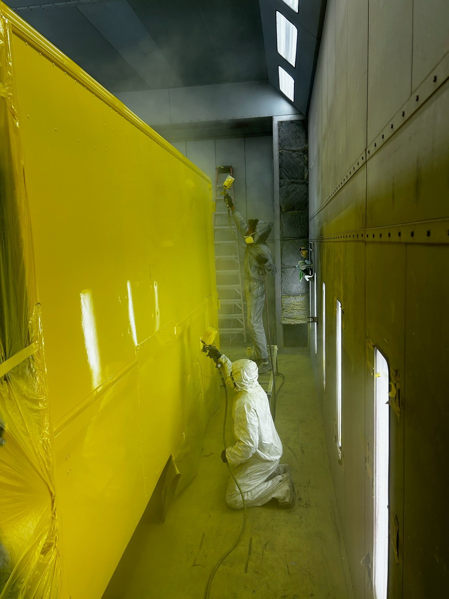 A person in a white protective suit spray-paints a large yellow panel inside an industrial paint booth.