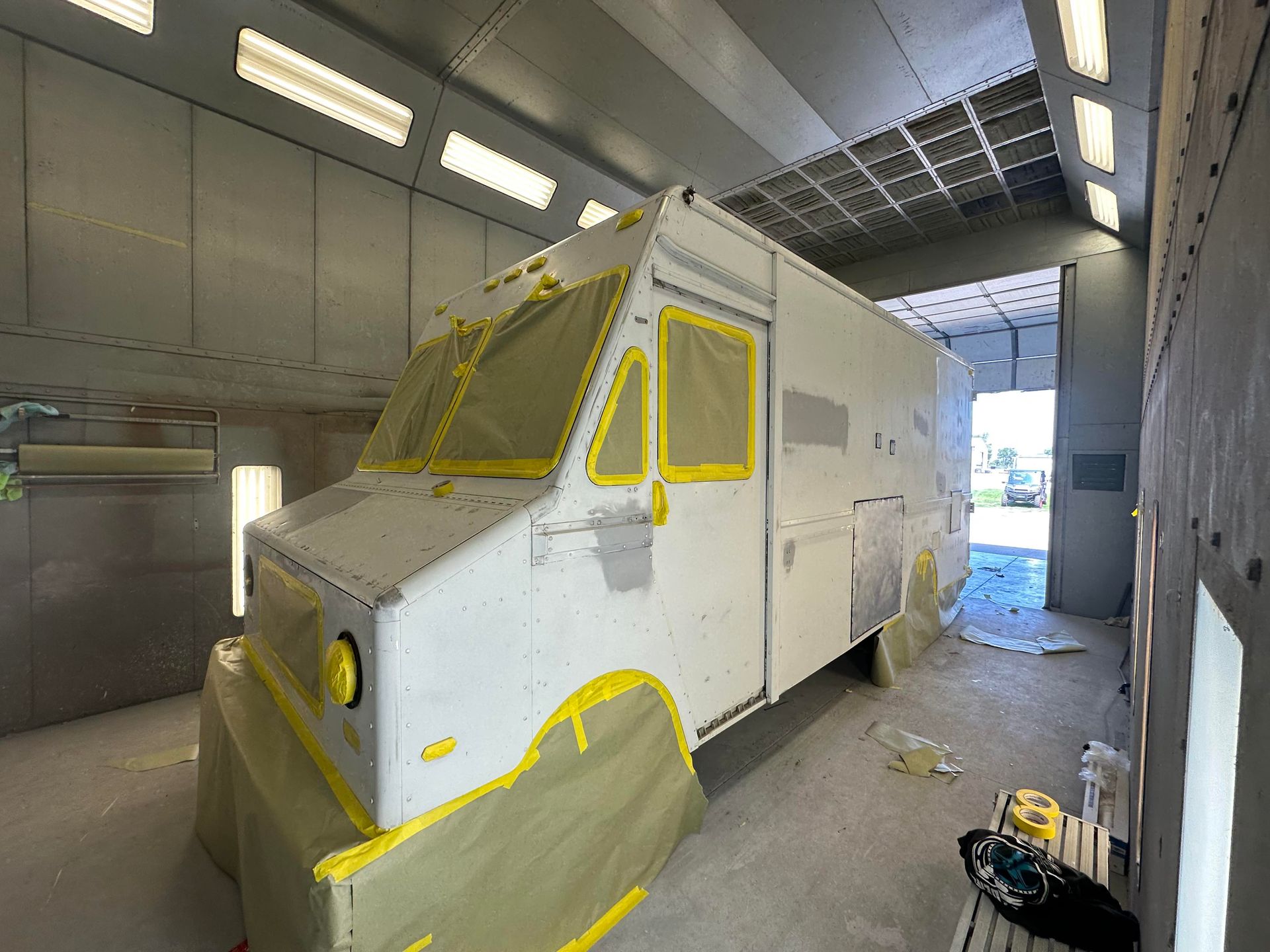 A white step van in an industrial paint booth with masked windows and wheels, partially sanded for repairs.