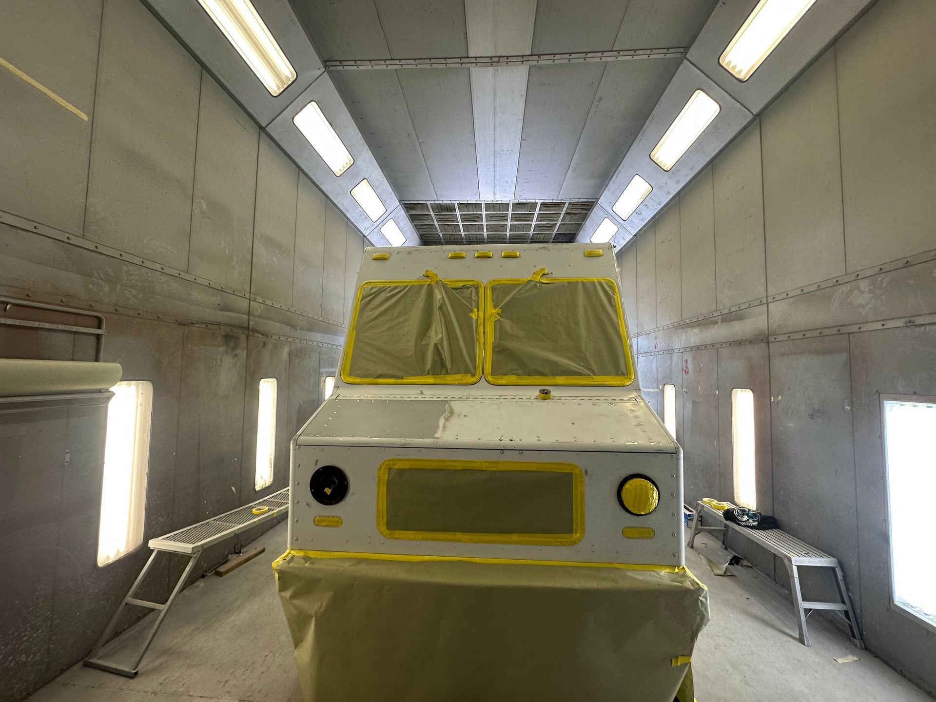 A white vehicle sits in a brightly lit industrial spray paint booth, with windows and lights masked by yellow tape.