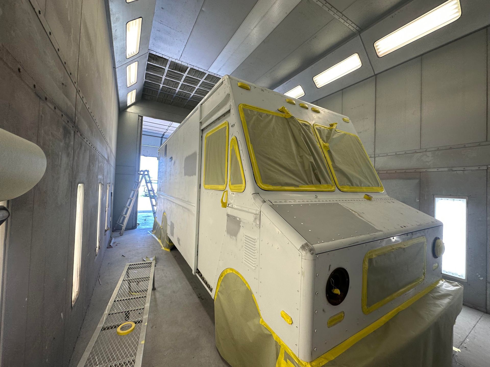 A white commercial delivery truck undergoing preparation for painting inside a brightly lit industrial spray booth.