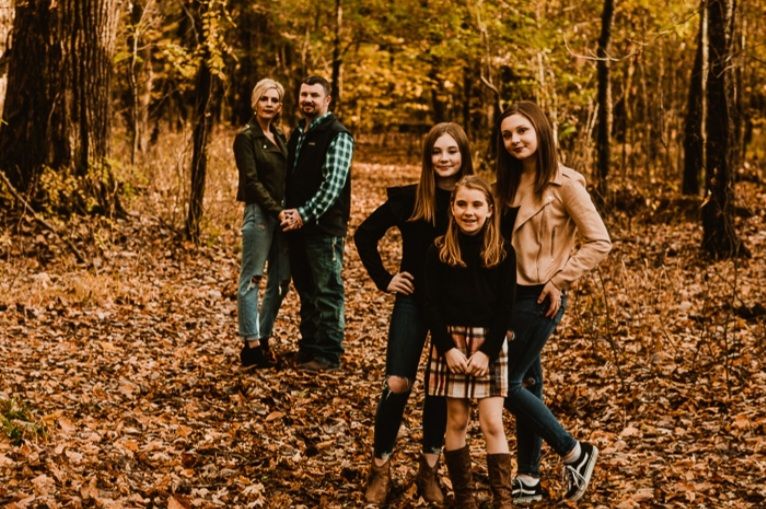 A group of five people poses in a forest with fallen autumn leaves, with two adults standing behind three children.