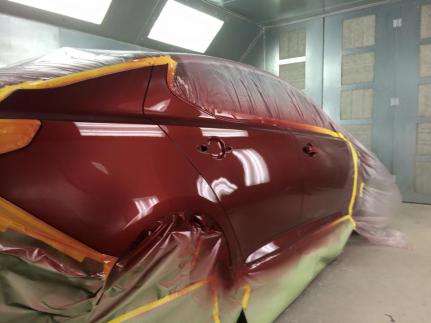 A maroon car side in a paint booth, partially masked with protective plastic and tape for a professional paint job.