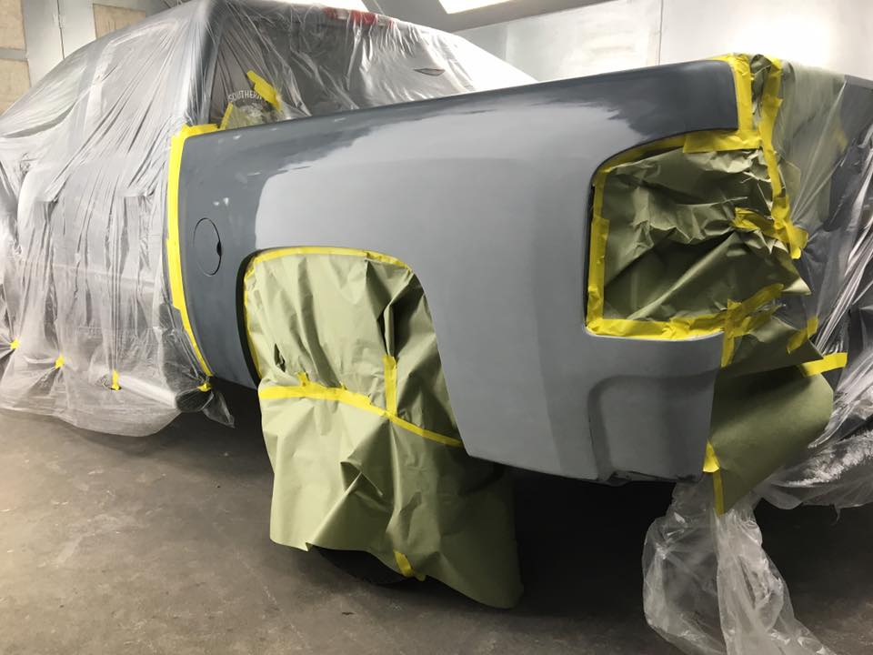 A grey truck bed in a paint booth, masked with tape and plastic for automotive repair.