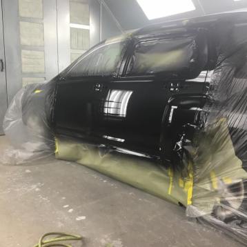 A black car being painted in a professional spray booth, with parts masked off by yellow tape and protective plastic.