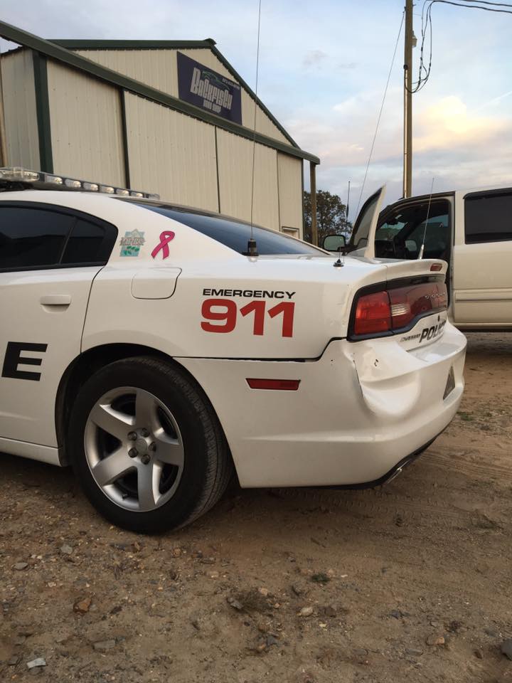 A white police cruiser with a damaged rear bumper parked on a gravel lot in front of a metal building.