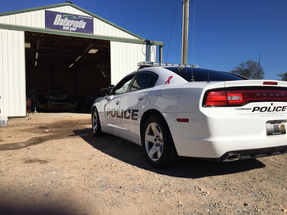 A white Dodge Charger police car parked outside a metal garage building under a clear blue sky.