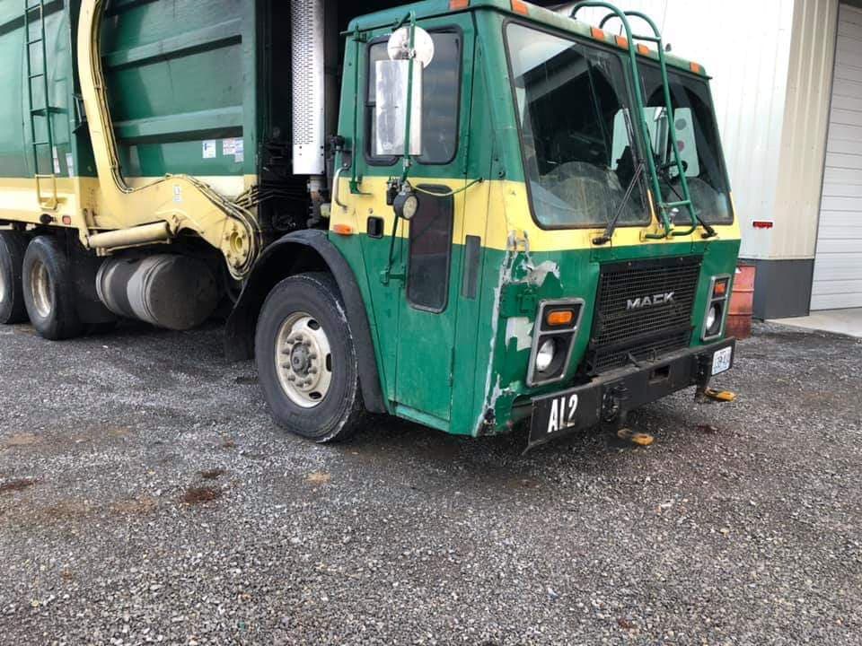 A green and yellow Mack garbage truck parked on a gravel surface.