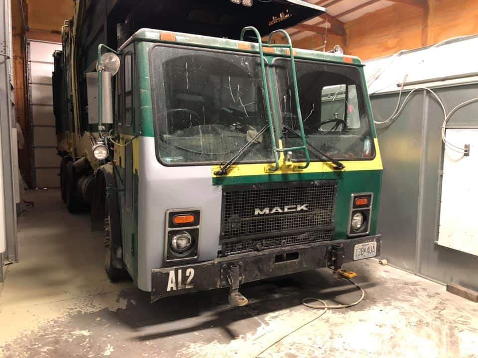 A Mack garbage truck with a partially painted green and grey front cab parked inside a garage.