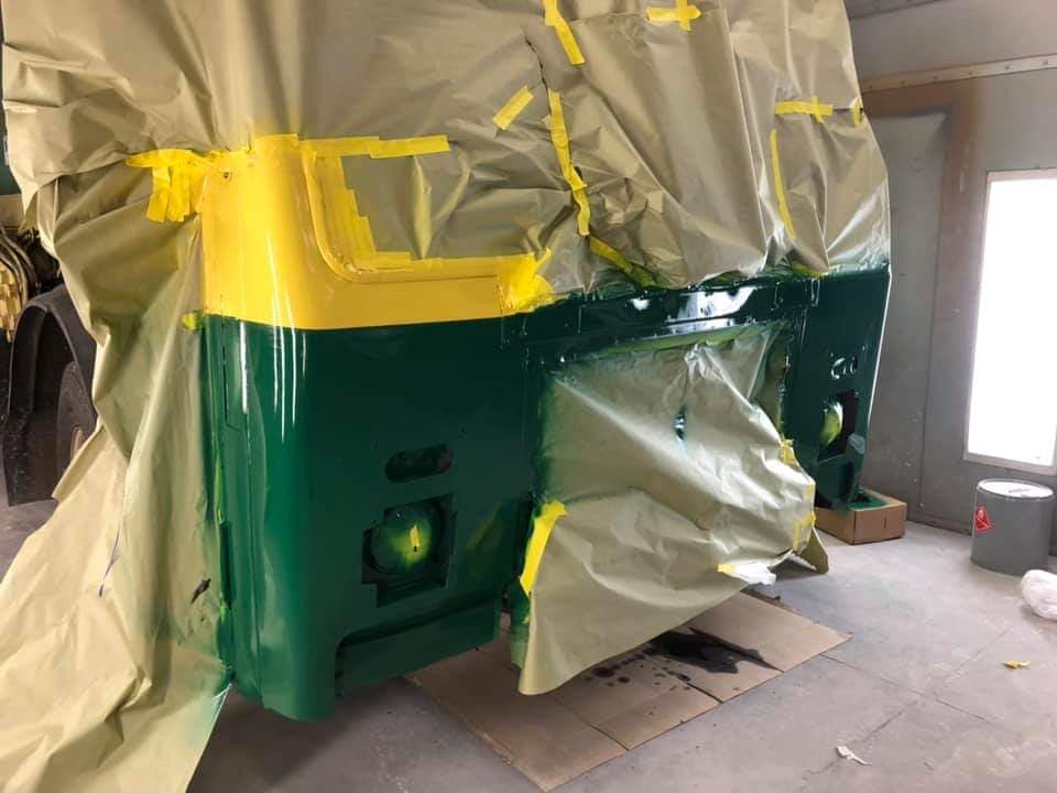A vehicle part in a workshop painted with a dark green lower section and a bright yellow upper, covered in masking paper.