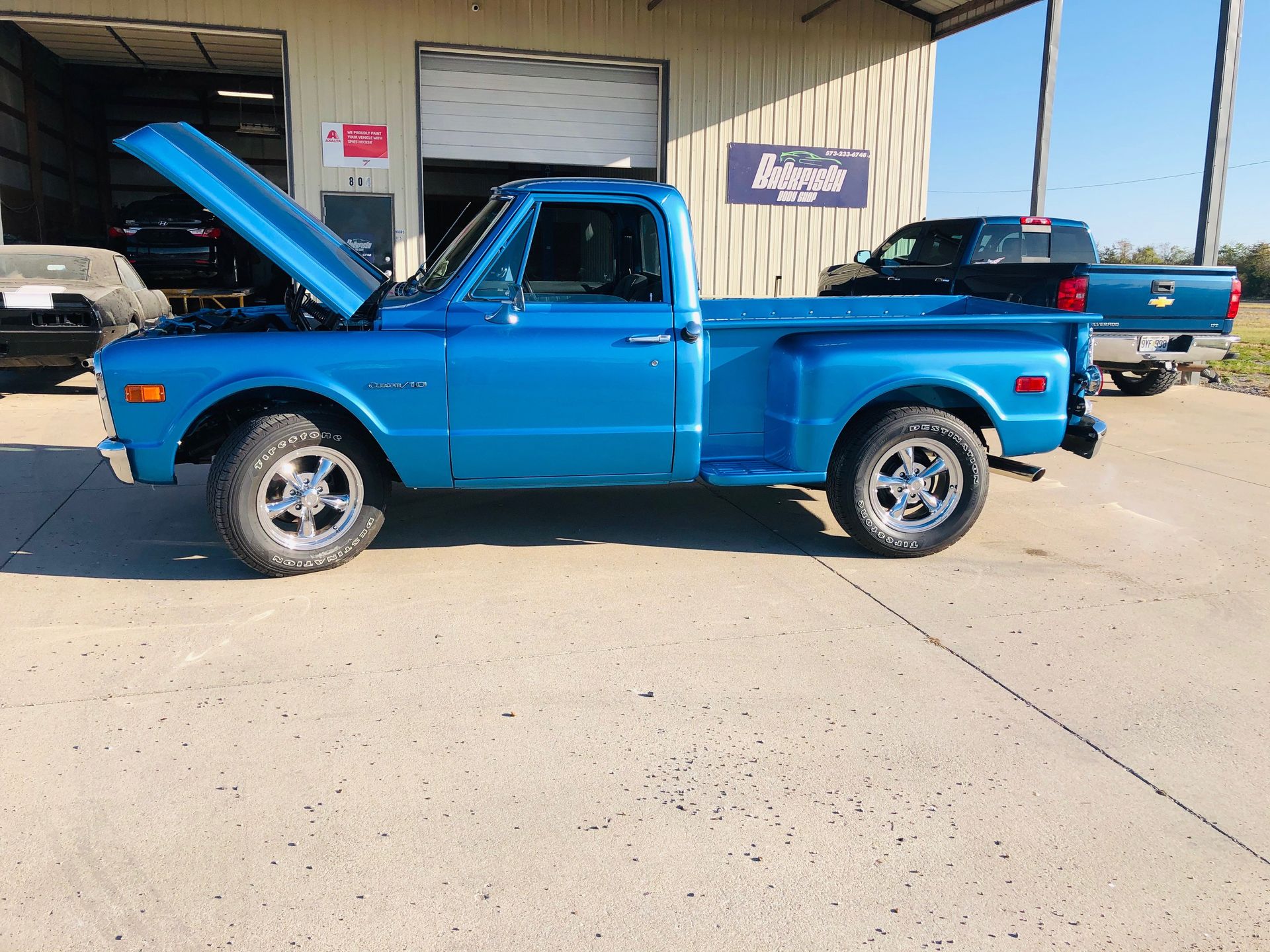A bright blue vintage Chevrolet C10 pickup truck with an open hood parked outside a garage on a sunny day.