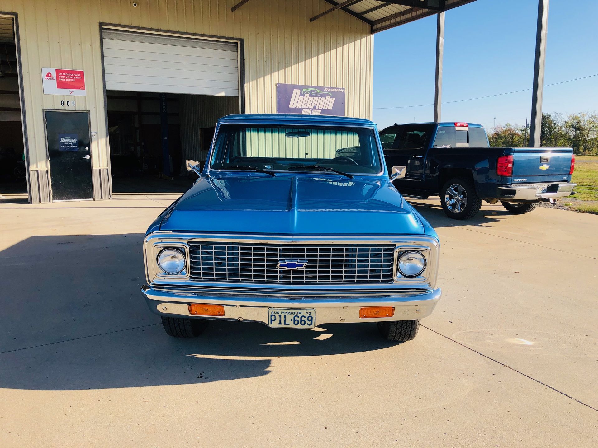 A bright blue vintage Chevrolet truck parked in front of a metal warehouse, with a dark blue pickup truck in the rear.