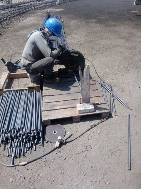 Un trabajador con casco azul se agacha junto a unas varillas metálicas y herramientas sobre un palé de madera al aire libre.