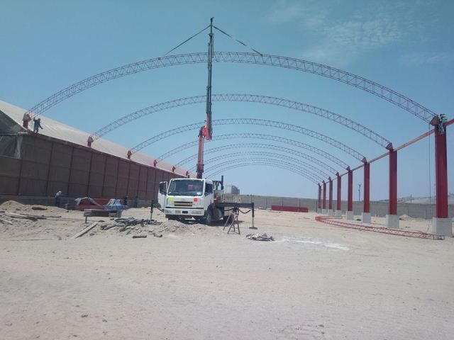Obra en construcción con un camión blanco, arcos de acero rojos y un muro de hormigón bajo un cielo despejado.