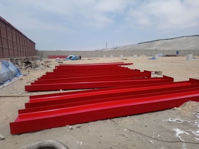 Vigas de acero rojo tendidas sobre terreno arenoso en una obra de construcción bajo un cielo nublado.