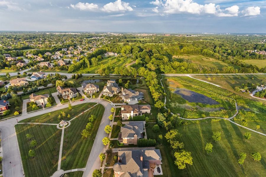 Aerial view of a suburban neighborhood with houses, curving roads, green fields, and a distant city skyline.