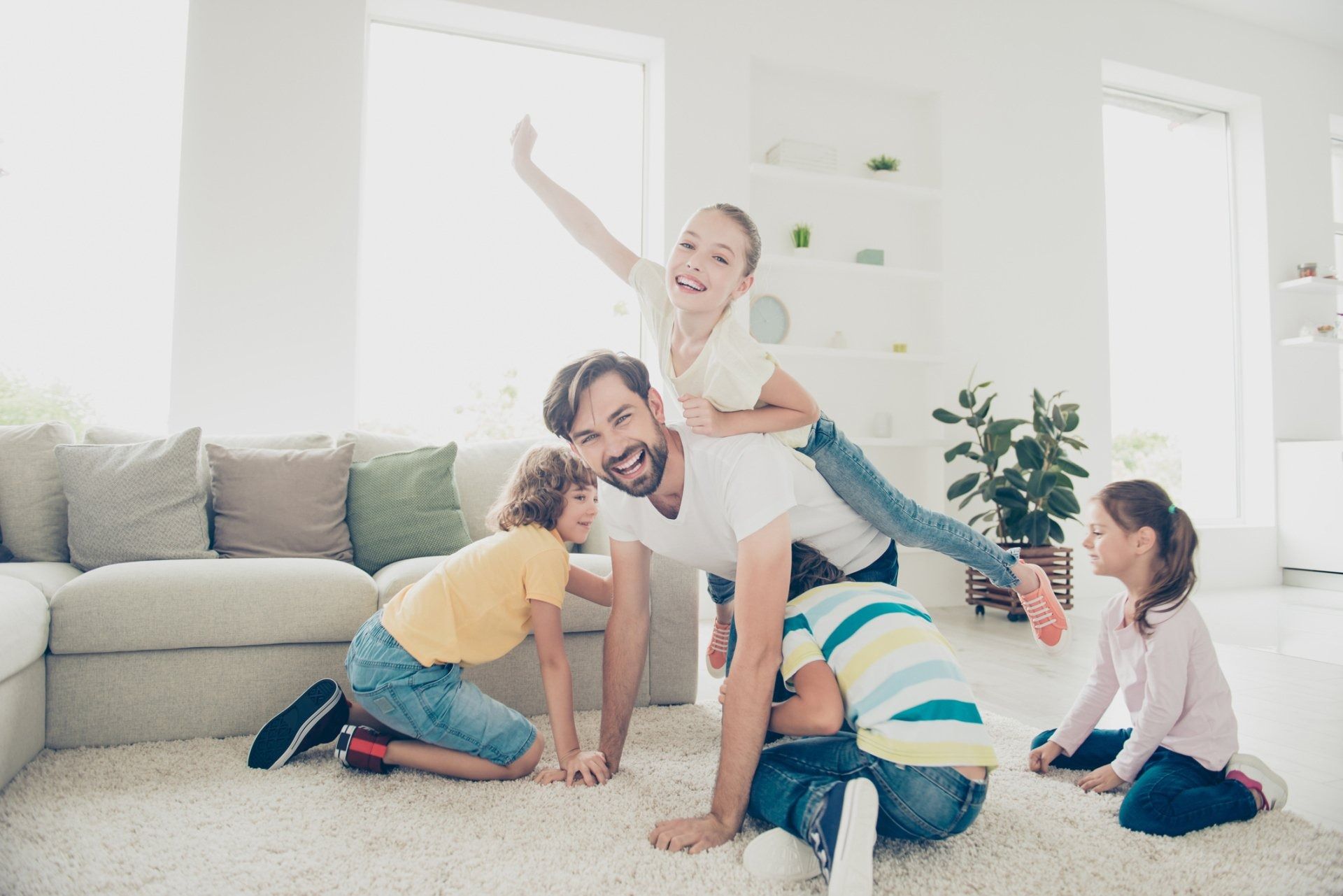 Family playing and stacking on a couch in a bright living room, with a child cheering on top.