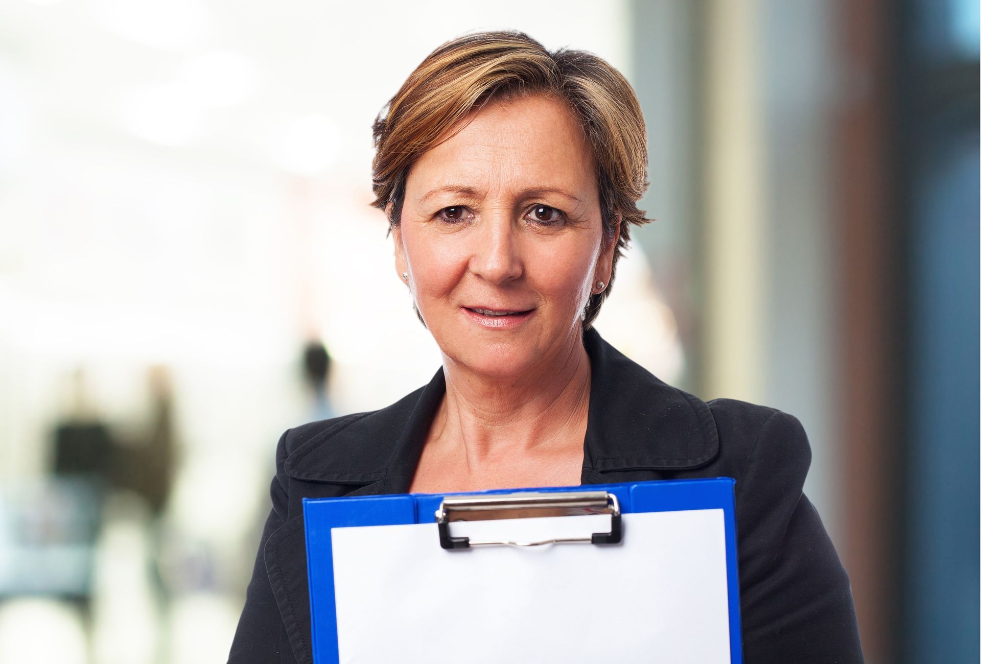 Woman holding a blue clipboard in an office setting, looking at the camera.