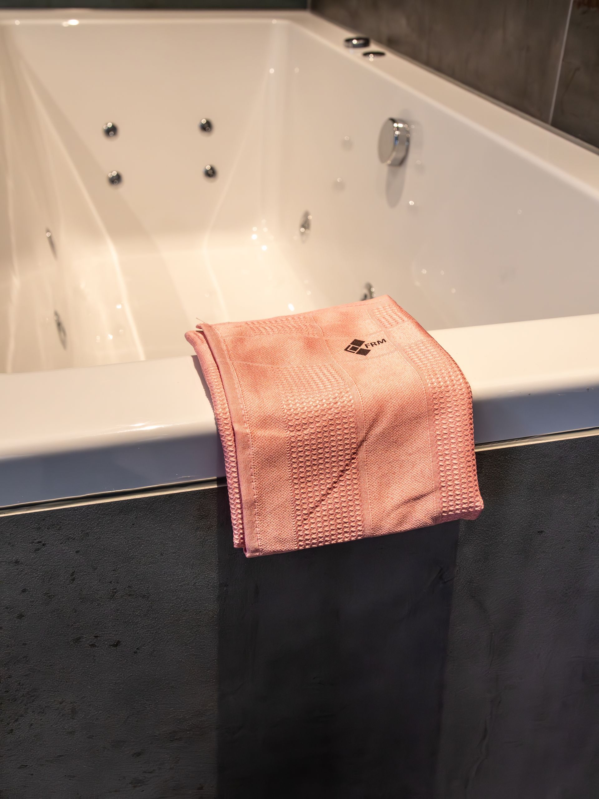 Een roze, gestructureerde microvezeldoek is over de rand van een wit bubbelbad met donkergrijze tegelwanden gevouwen.