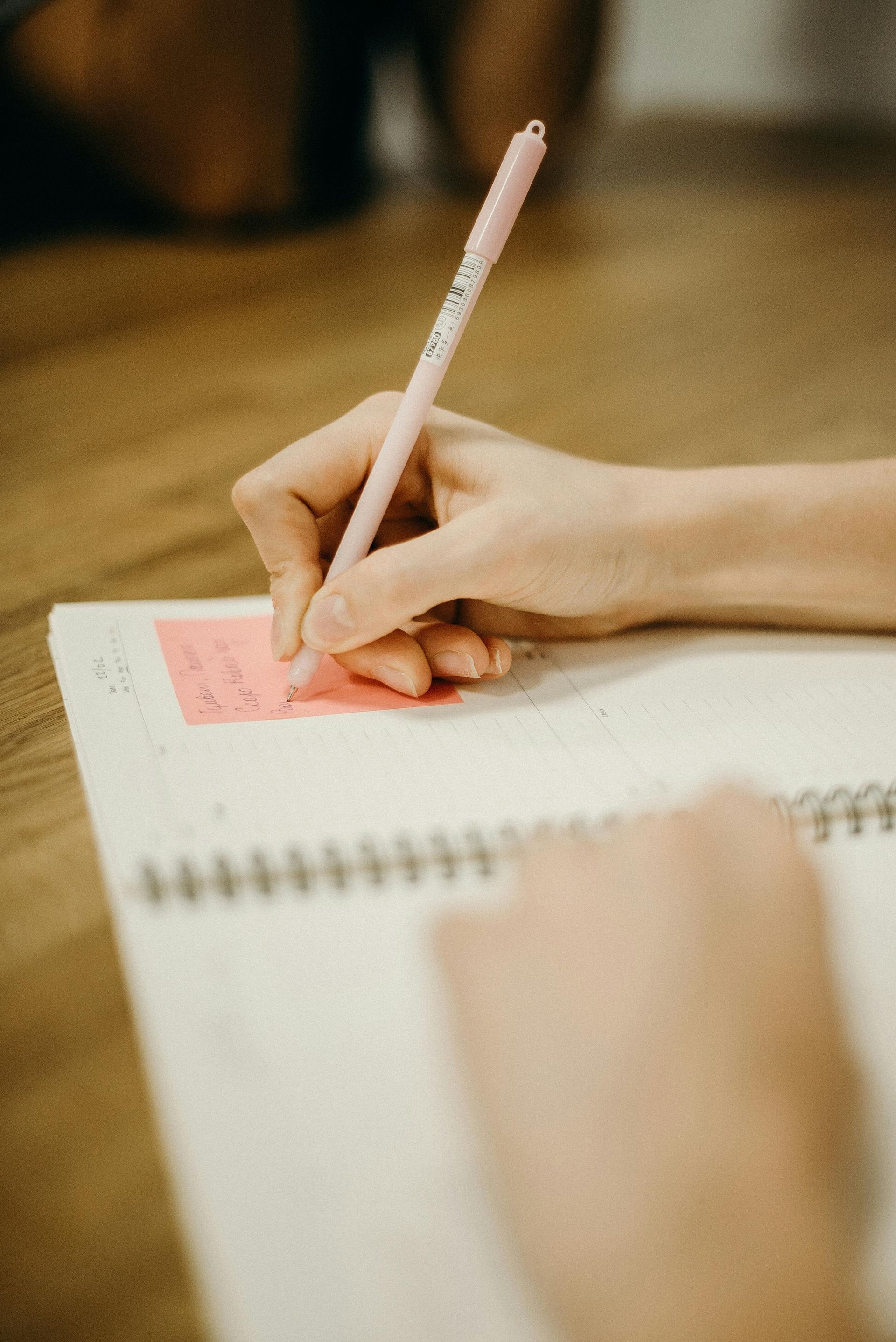 Een hand schrijft met een lichtroze pen op een klein roze plakbriefje dat vastzit aan een spiraalblok op een houten tafel.