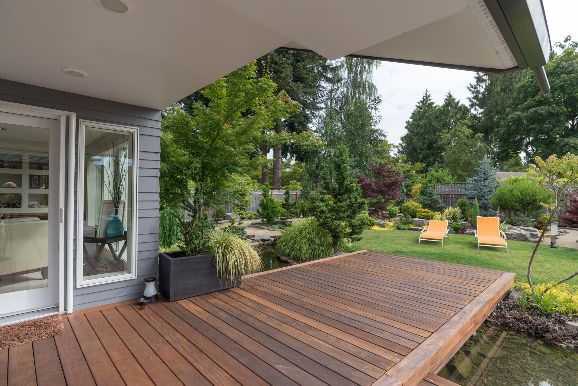 Wooden deck with water feature, lush green backyard, and modern house. Two orange chairs on the grass.