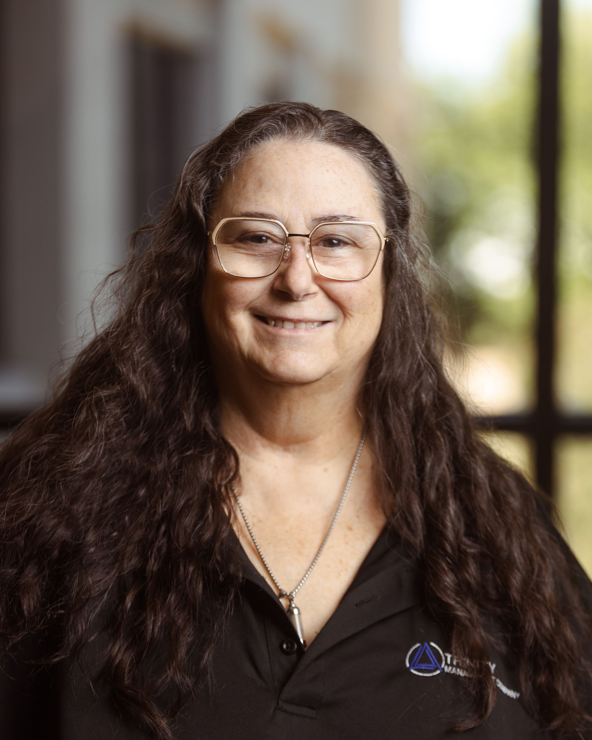 Smiling person with long curly hair and glasses, wearing a black shirt indoors