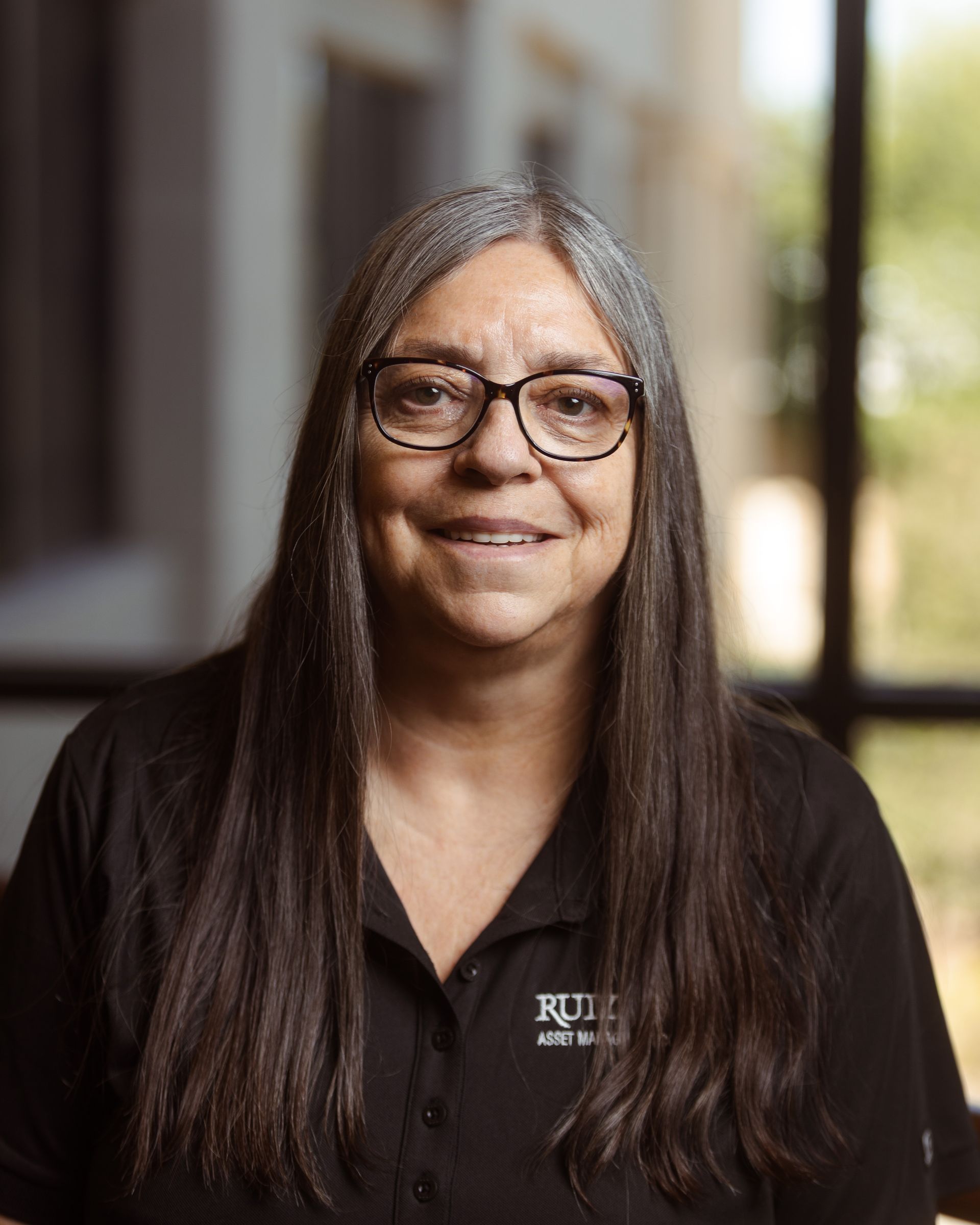 Smiling person with long dark hair and glasses in a black shirt, seated indoors with a blurred background.