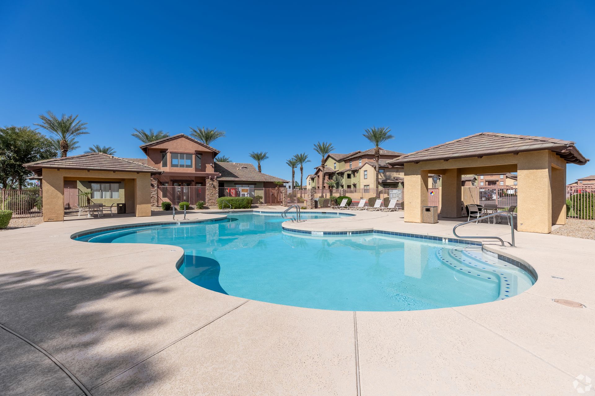 Desert resort pool with turquoise water, beige cabanas, and tan buildings under a clear blue sky