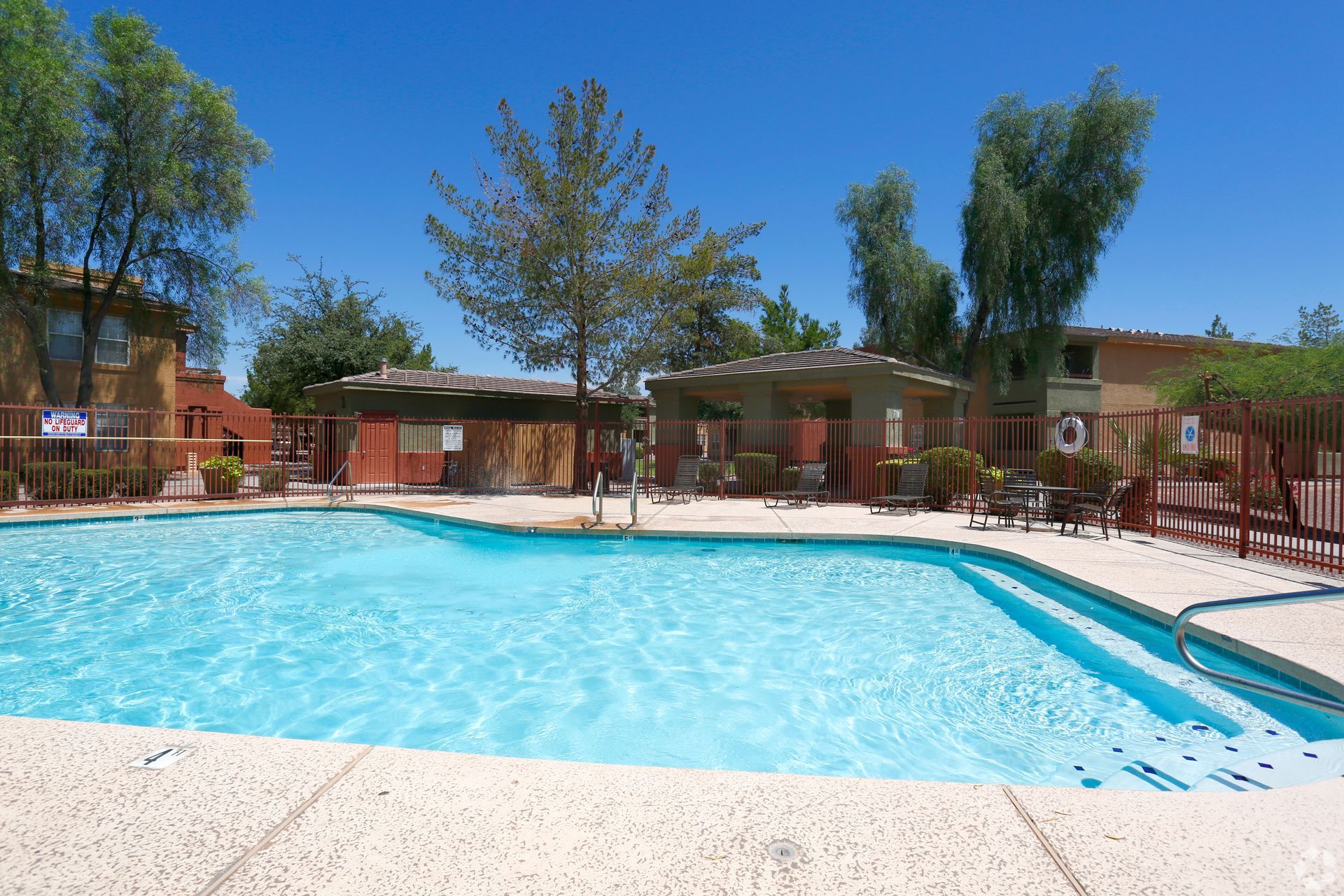 Outdoor swimming pool with clear blue water, lounge chairs, and trees under a sunny sky