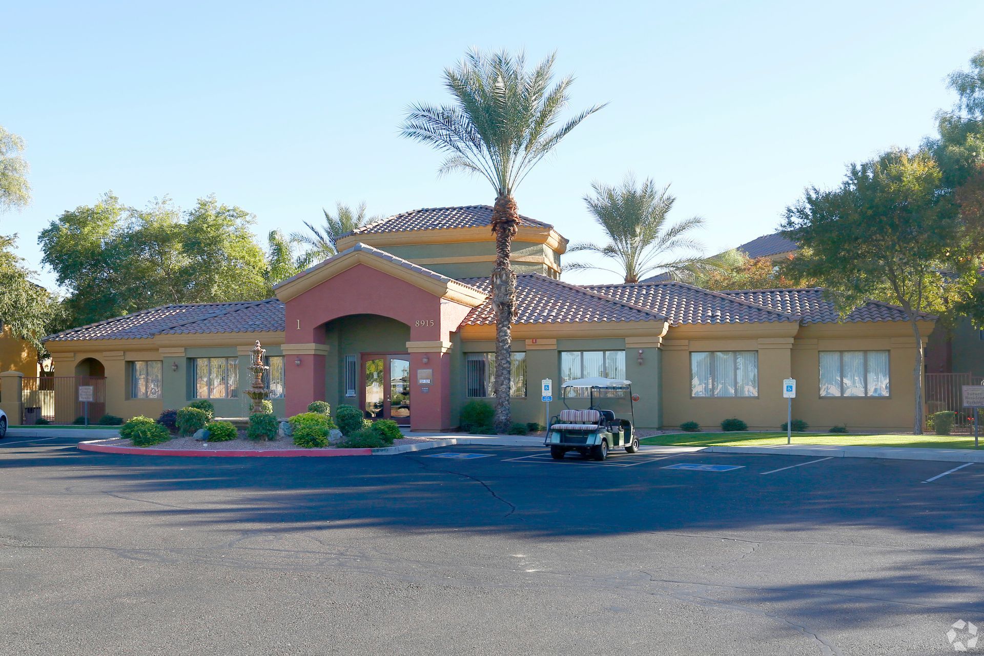 Single-story beige building with a tiled roof, palm trees, and a parked golf cart in front.