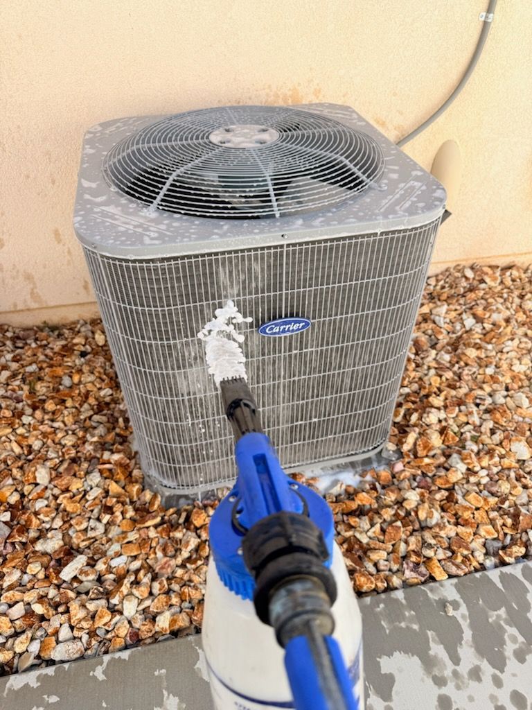 Pressure washer spraying a dirty outdoor air conditioner unit on gravel beside a wall