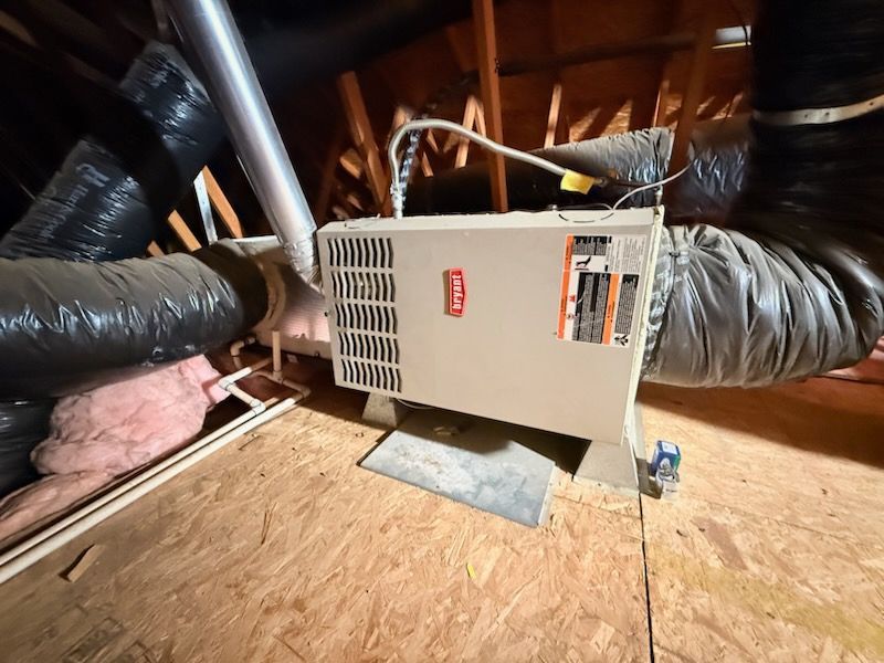 Furnace unit in an attic surrounded by black insulated ducts and exposed wooden rafters.
